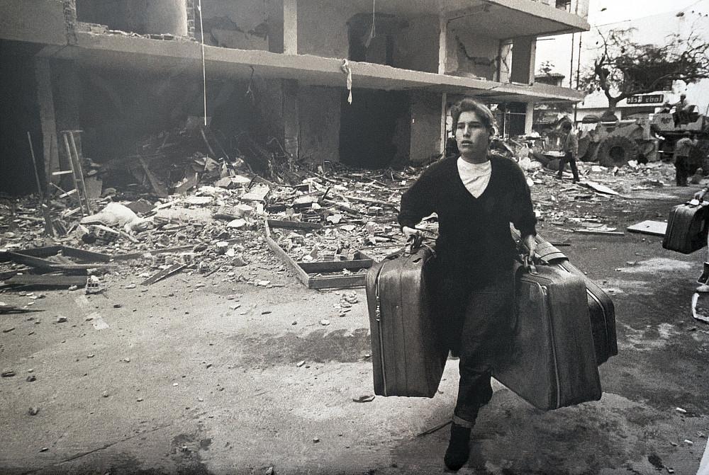 A person runs with three bags of luggage through ruined buildings.